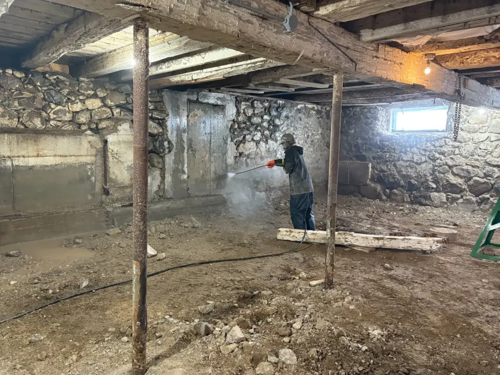 Professional interior barn foundation restoration showing deep pressure washing and surface preparation of an old fieldstone wall to ensure maximum shotcrete adhesion.